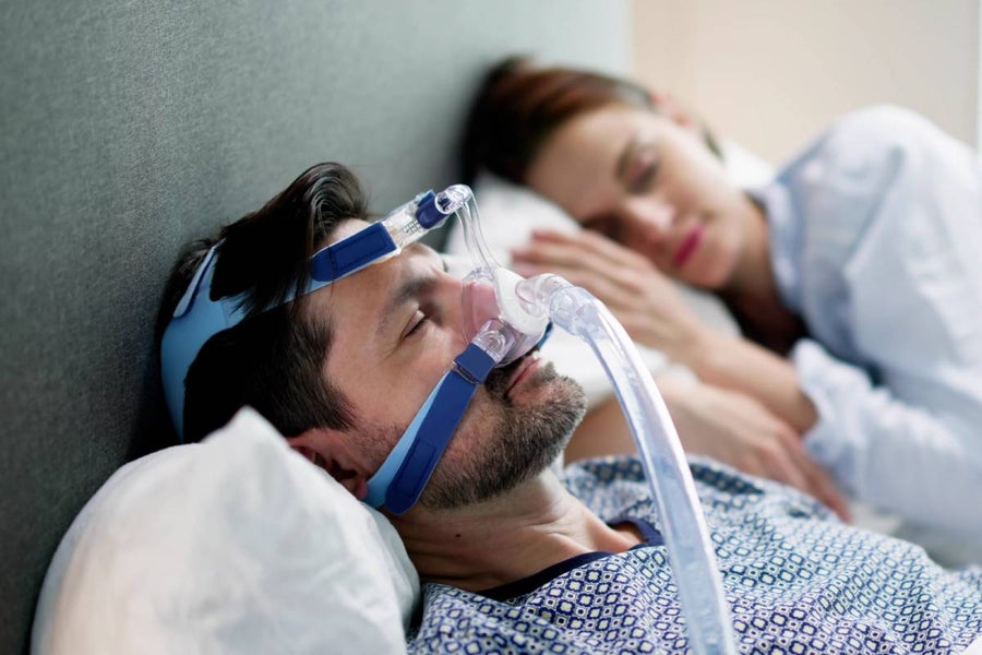 Man sleeping with a CPAP mask on, beside a blurred woman also sleeping.