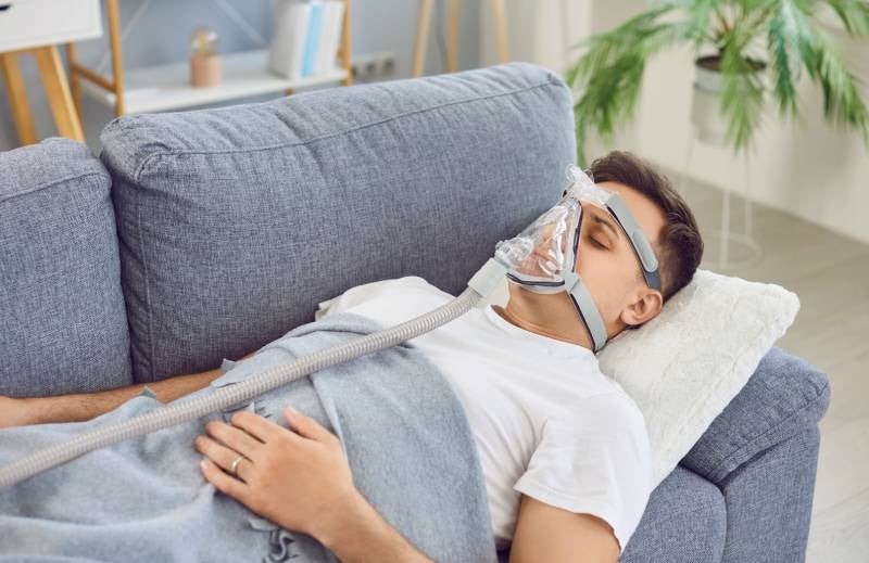 A man sleeping on a gray couch wearing a full-face CPAP mask with a tube extending down his body.