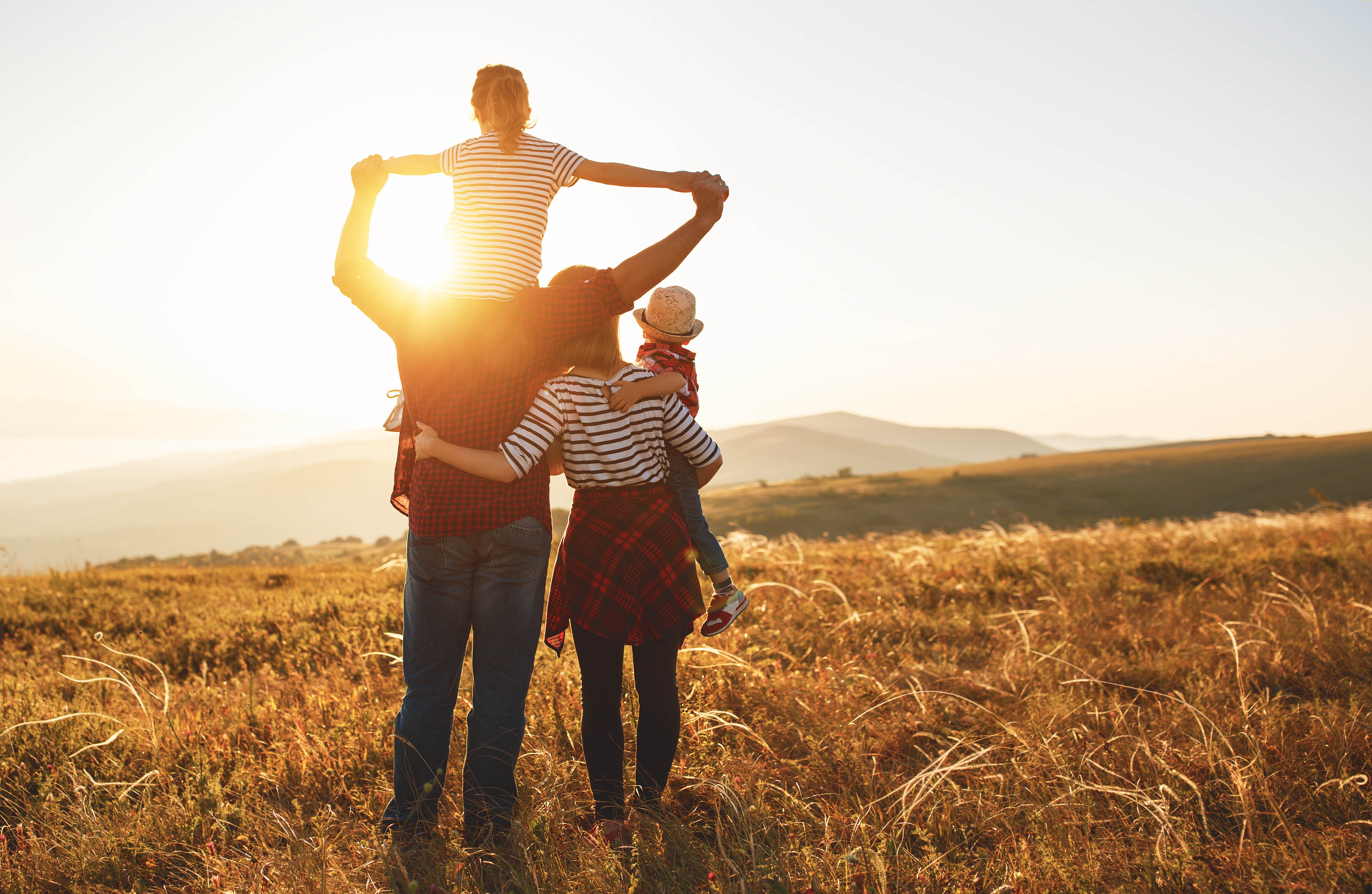 A family of four stands in a sun-drenched field, children on parents' shoulders, facing away towards mountains.