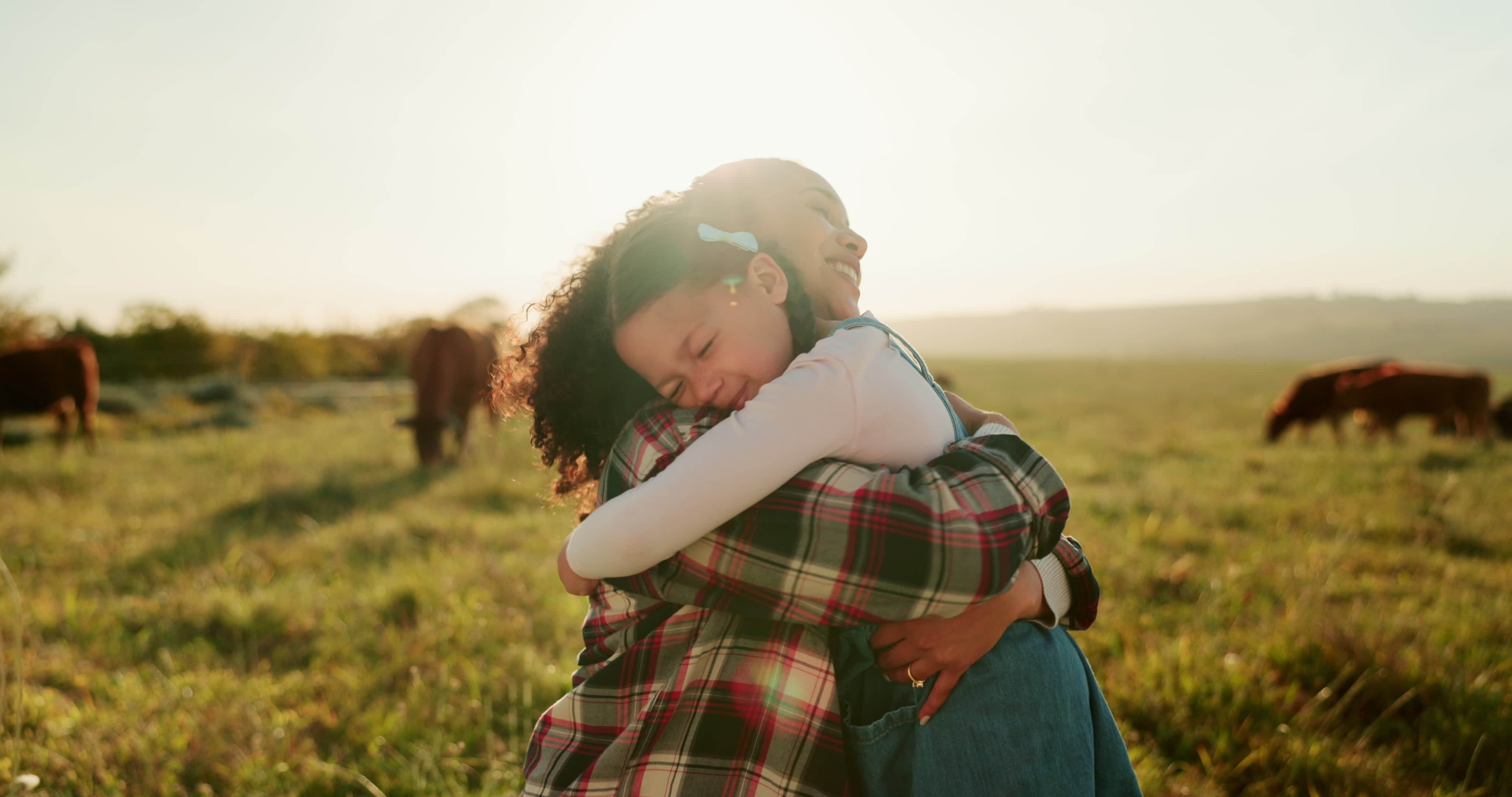 Happy Black mother and daughter hugging in a sunny field with cattle grazing in the background.