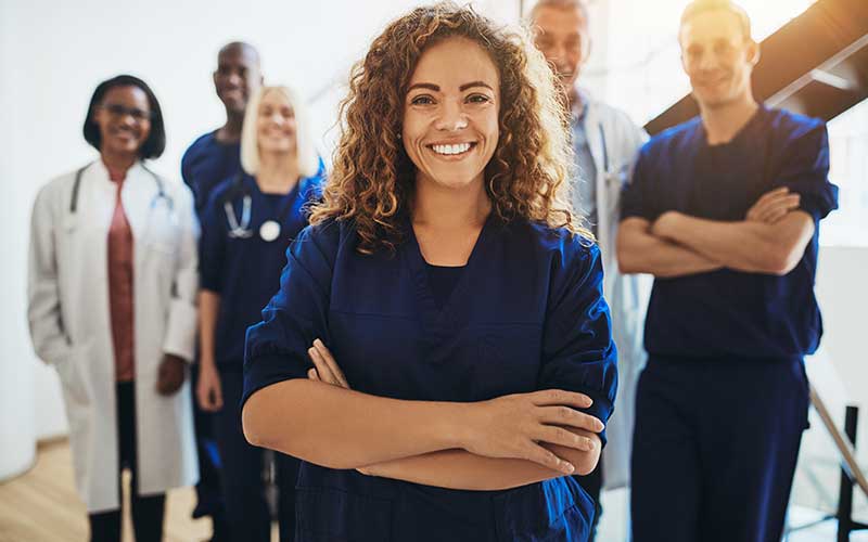 A smiling healthcare worker in scrubs stands confidently with colleagues in a modern medical setting.