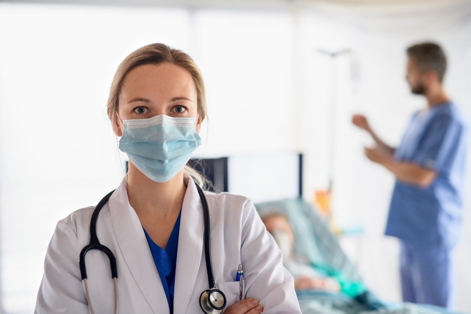 A healthcare worker wearing a mask and stethoscope looks directly at the viewer in a hospital setting.