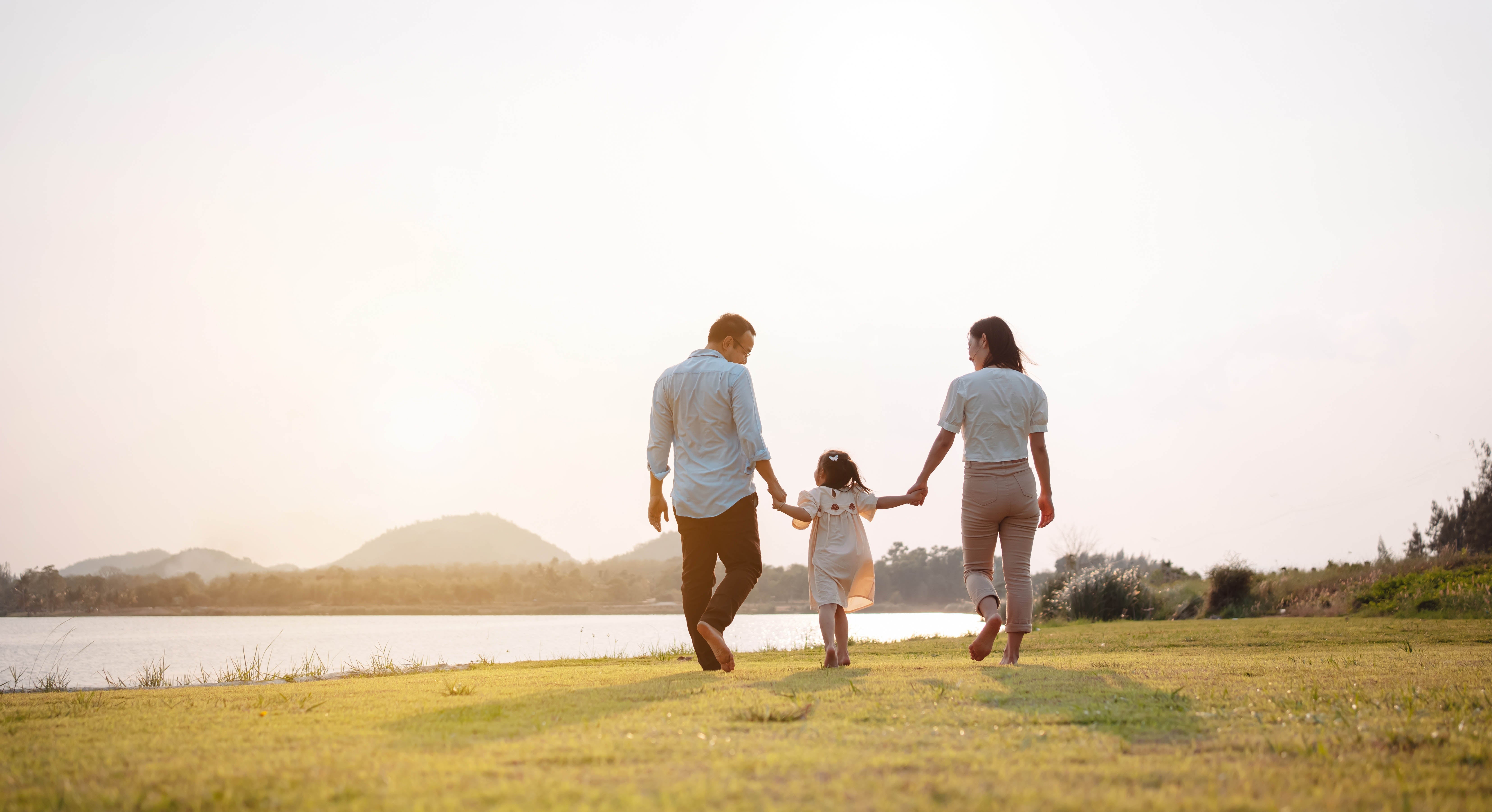 Full-length, rear view of a family of three holding hands as they walk barefoot on grass near a lake.